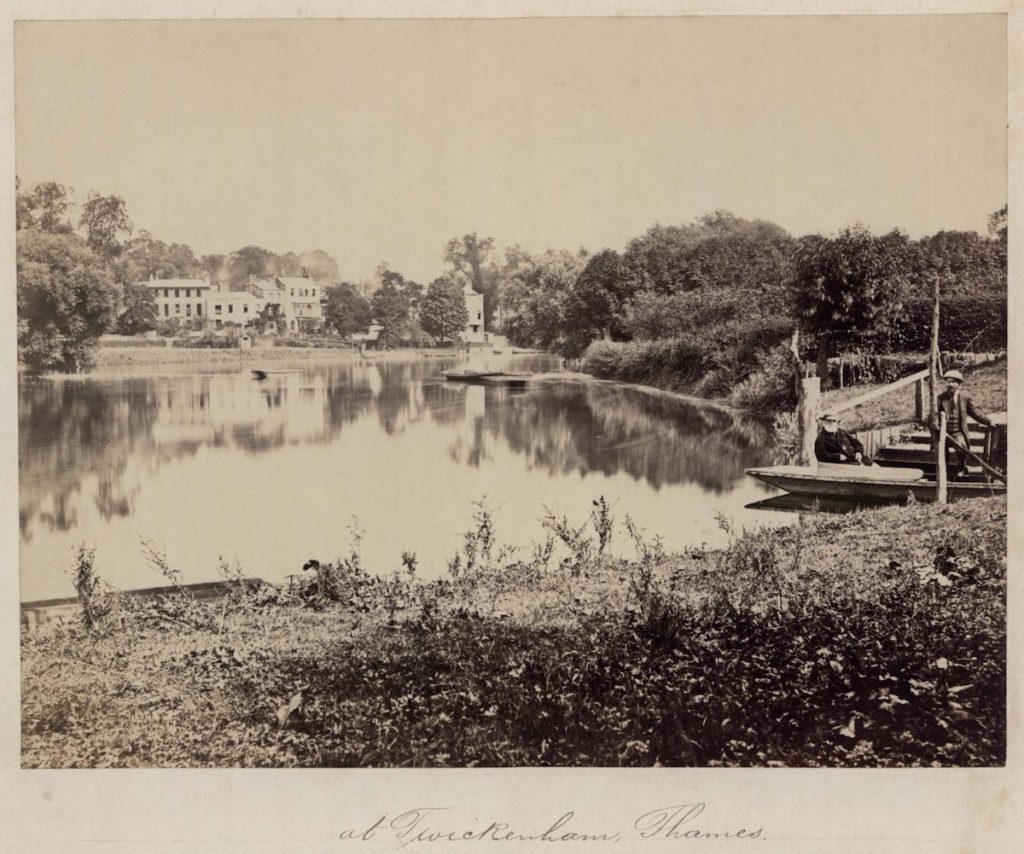 Sepia photo of a lake with large country house in the background.