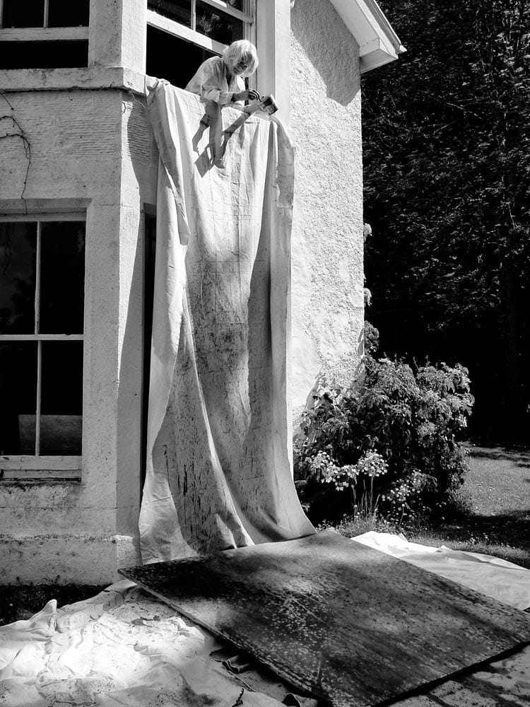 Jane leaning out of a first floor window, wielding paint can and brush, splashing paint onto a large canvas below. Sheets protect the walls of the building, and ground surrounding the area.