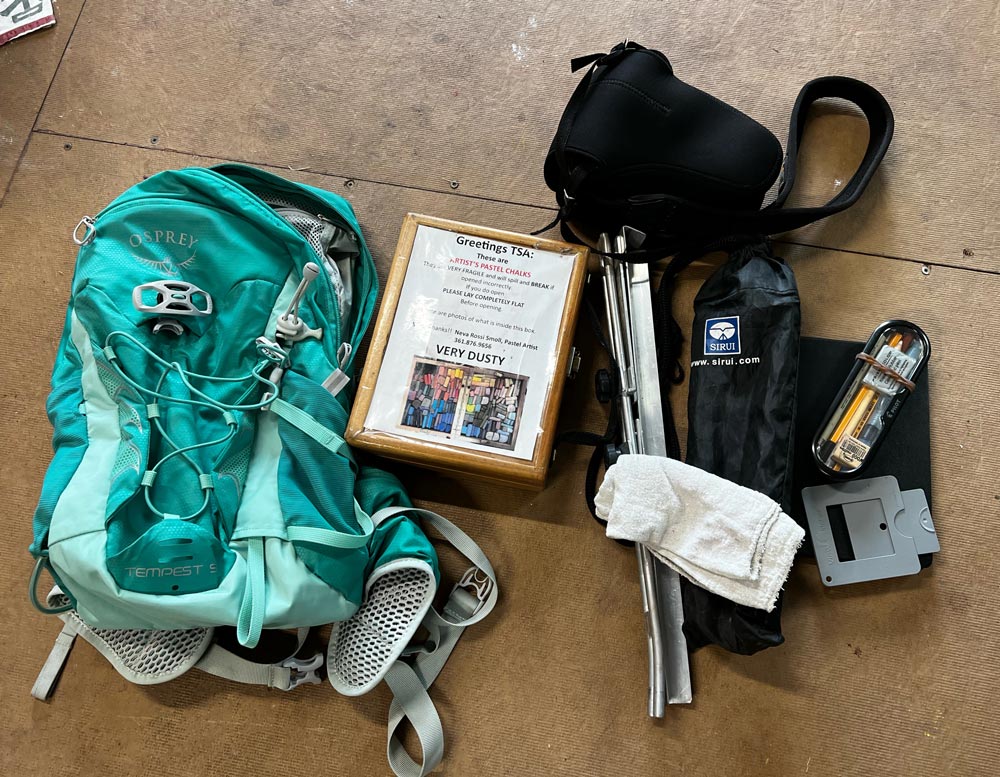 Backpack, pastel box, tripod, sketchbook and camera laid out on the floor.