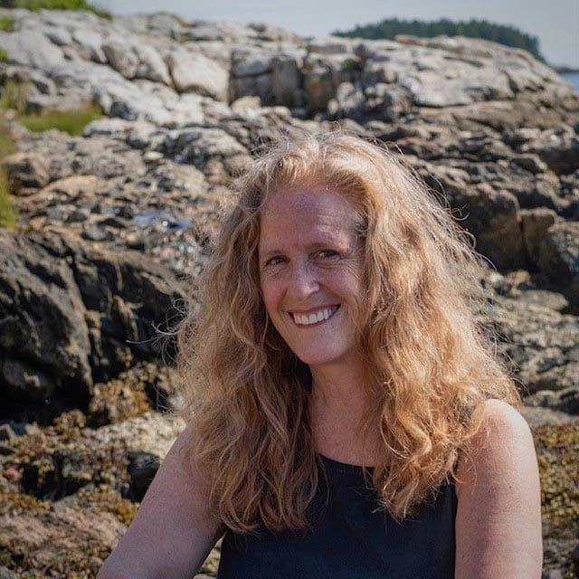 Lyn Asselta sits on a rocky shoreline side lit by the bright sunshine.