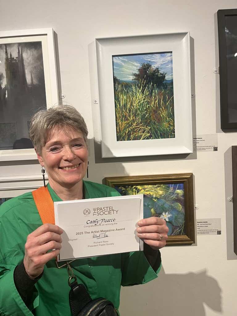 Cathy Pearce stands next to her painting 'Bright and Early', while holding her 'The Artist Magazine Award 2025' certificate.